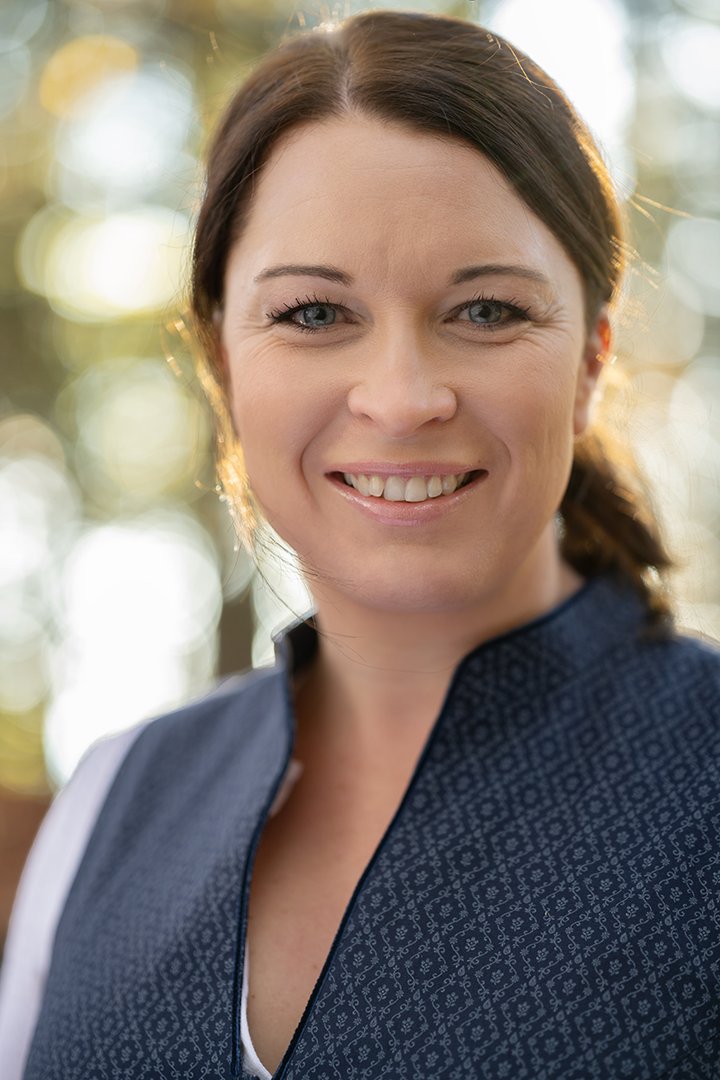 Foto: Tourismusverband Hausruckwald/Thomas Koller: Team_Bernadette Nagl Portraitfoto von Hausruckwald-Teammitglied Bernadette Nagl. Dunkelbraune Haare zu einem Pferdeschwarz gebunden, dunkelblaues Dirndl mit Stehkragen und weißer, schlichter Bluse. Sie lächelt und schaut direkt in die Kamera.