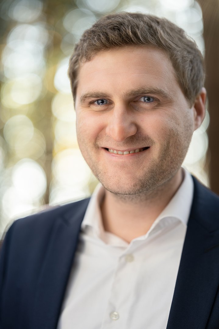 Foto: Tourismusverband Hausruckwald/Thomas Koller: Geschäftsführer_Elias Gavino-Schlager Portraitfoto von Hausruckwald-Geschäftsführer Elias Gavino-Schlager. Dunkelblonde, kurze Haare, dunkelblaue Anzugjacke und weißes Hemd, oberster Knopf offen. Er lächelt und schaut direkt in die Kamera.